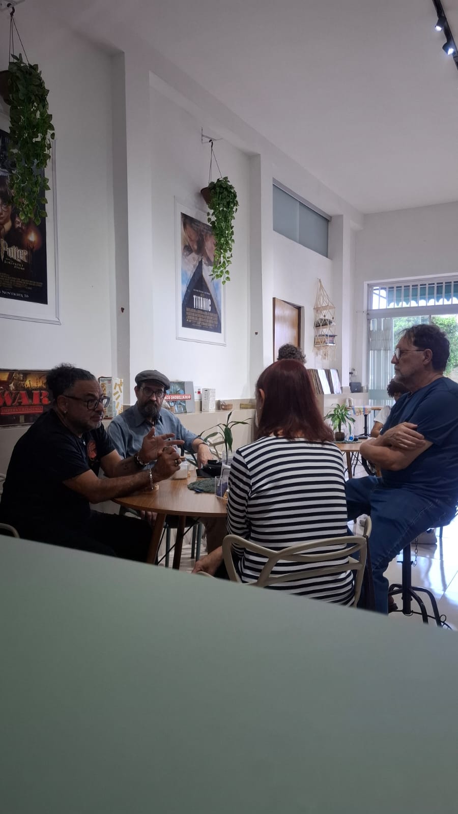 Grupo de clientes reunido numa mesa do Sebo Araxá, conversando sob pôsteres de filme e plantas penduradas.